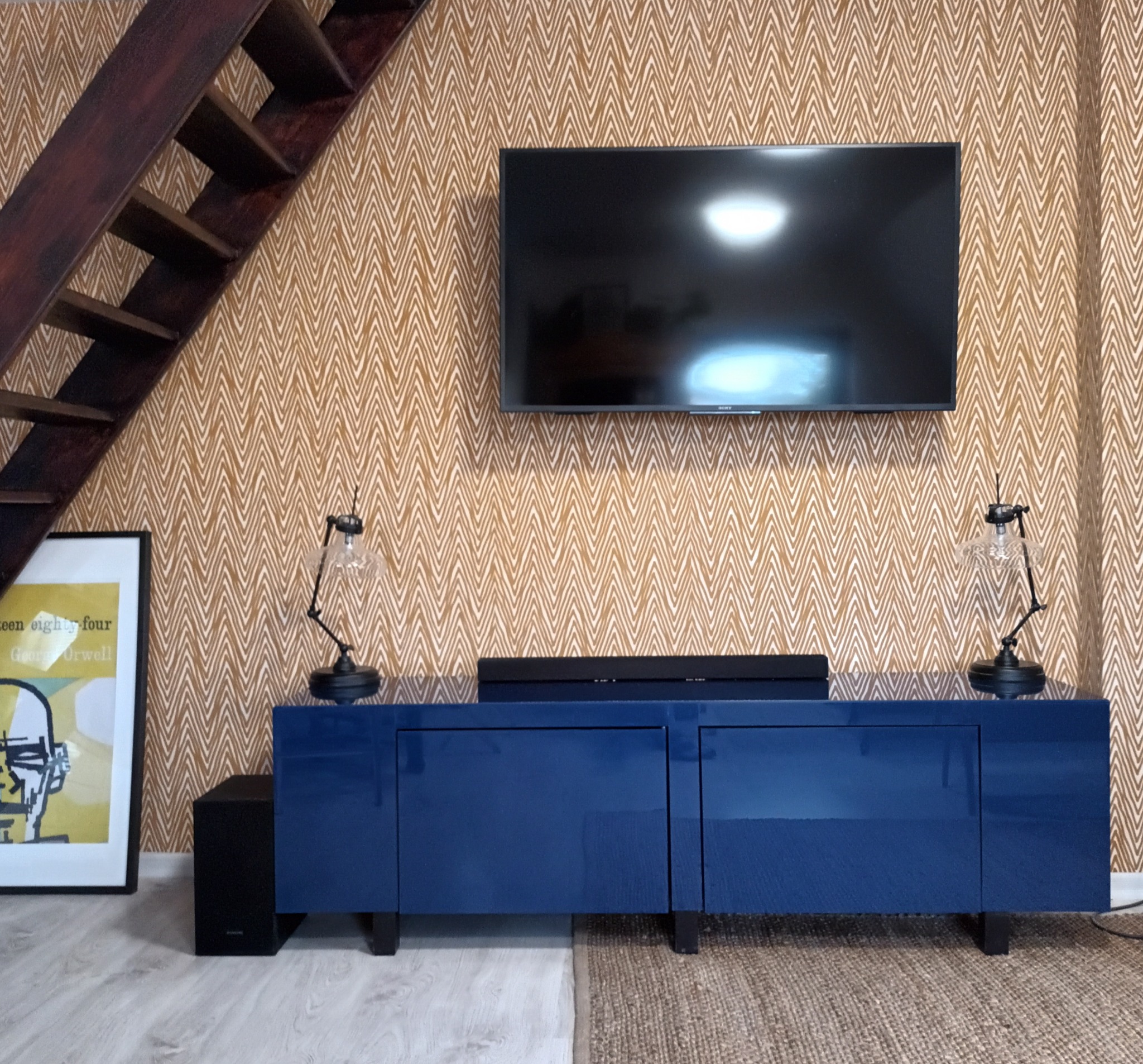 Niebieska szafka RTV z połyskiem pod wiszącym telewizorem, lampki nocne z kloszami, plakat i drewniane schody na tle ściany w zygzakowate wzory.
