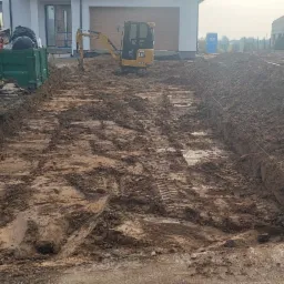 Przygotowanie terenu pod budowę w Lublinie: wykopane rowy, mała koparka CAT, hałda ziemi i kontener na śmieci budowlane w tle.