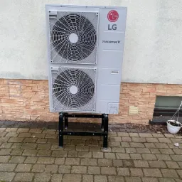 Zewnętrzna jednostka pompy ciepła LG Therma V na metalowym stelażu, ustawiona na kostce brukowej przed budynkiem z fragmentem ceglanej elewacji i białym murem. Pusta doniczka po prawej stronie.