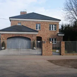 Dwukondygnacyjny dom z cegły klinkierowej z garażem z łukowym wjazdem, szarą bramą i numerem 33 nad wejściem, otoczony metalowym ogrodzeniem na ceglanej podmurówce.