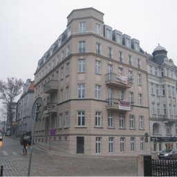 BUD-OWY SPÓŁKA Z OGRANICZONĄ ODPOWIEDZIALNOŚCIĄ - Kamienica po remoncie elewacji w miejskiej zabudowie, widoczna z perspektywy ulicy wyłożonej kostką, z banerem reklamowym na balkonie.