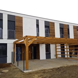 BUD-OWY SPÓŁKA Z OGRANICZONĄ ODPOWIEDZIALNOŚCIĄ - Nowoczesny dom szeregowy z drewnianą pergolą nad miejscem parkingowym, elewacja w kolorze białym i drewnopodobnym, okna z roletami zewnętrznymi.