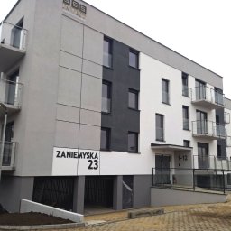 BUD-OWY SPÓŁKA Z OGRANICZONĄ ODPOWIEDZIALNOŚCIĄ - Nowoczesny budynek mieszkalny z balkonami, elewacją w odcieniach szarości i bieli, widoczny adres 'Zaniemyska 23' oraz żółty samochód dostawczy w tle.