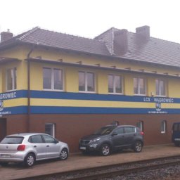 BUD-OWY SPÓŁKA Z OGRANICZONĄ ODPOWIEDZIALNOŚCIĄ - Odnowiony budynek stacji kolejowej z żółtą elewacją, brązowym cokołem i niebieskim pasem, z zaparkowanymi samochodami i torami kolejowymi w kadrze.