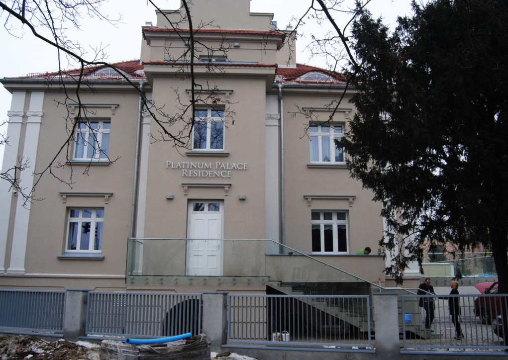 Elewacja budynku 'Platinum Palace Residence' w beżowej kolorystyce z białymi oknami, widoczny fragment ogrodzenia i drzewa