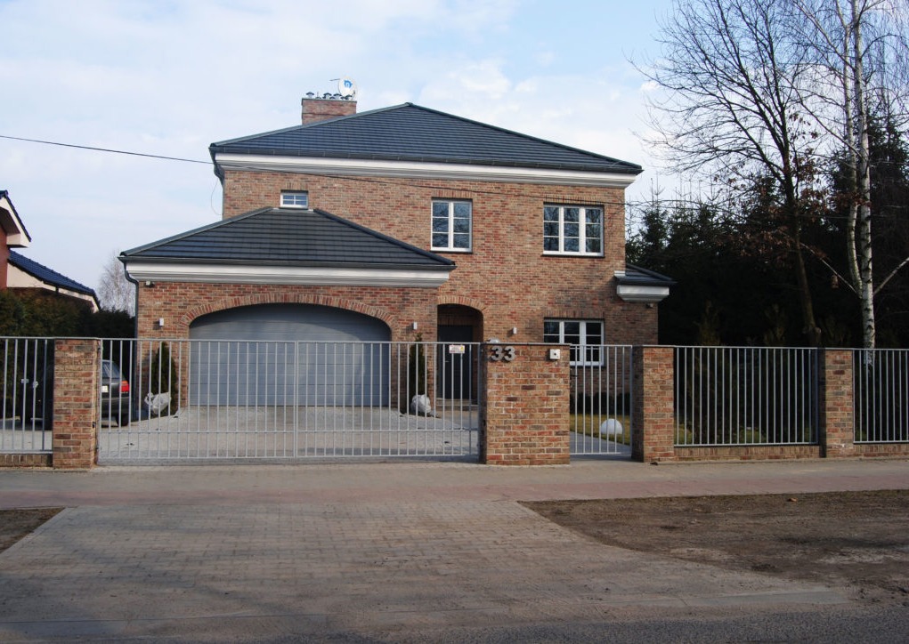 Dwukondygnacyjny dom z cegły klinkierowej z garażem z łukowym wjazdem, szarą bramą i numerem 33 nad wejściem, otoczony metalowym ogrodzeniem na ceglanej podmurówce.