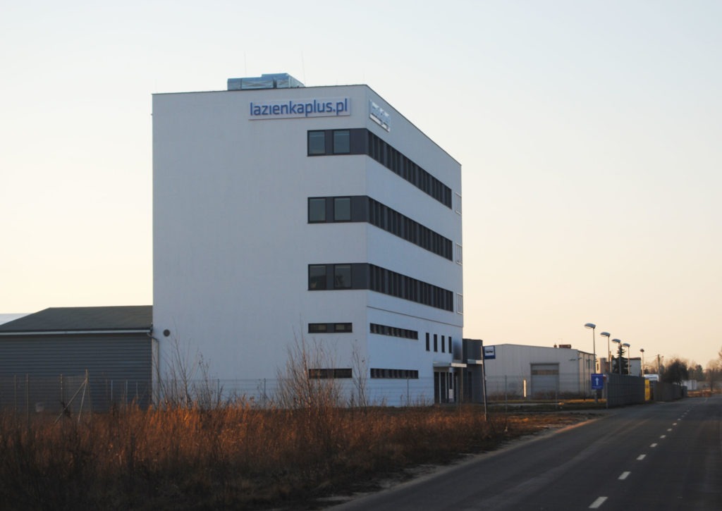 Biały, nowoczesny budynek biurowy z napisem lazienkaplus.pl, widoczny z perspektywy ulicy, z suchą trawą na pierwszym planie.