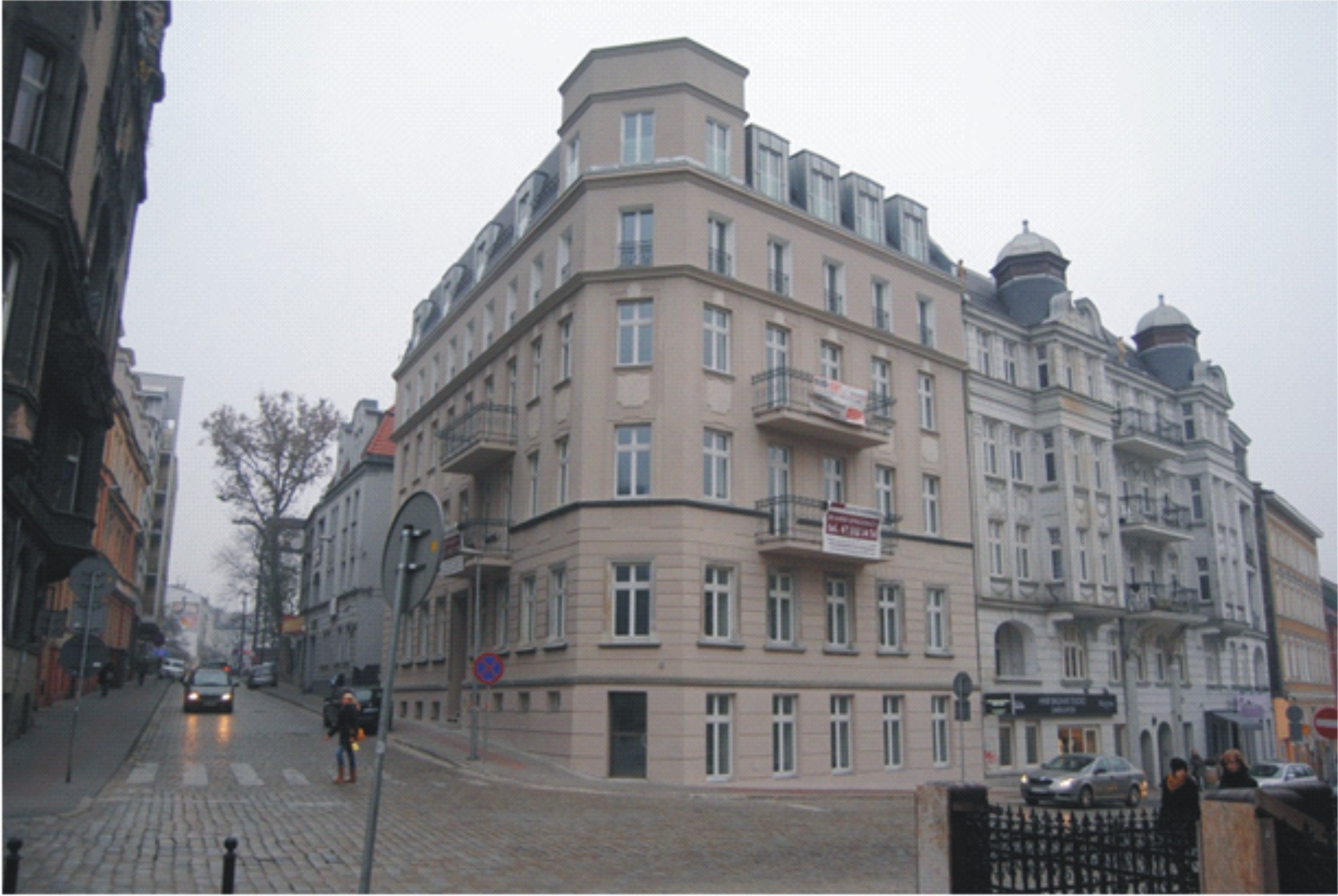 Kamienica po remoncie elewacji w miejskiej zabudowie, widoczna z perspektywy ulicy wyłożonej kostką, z banerem reklamowym na balkonie.