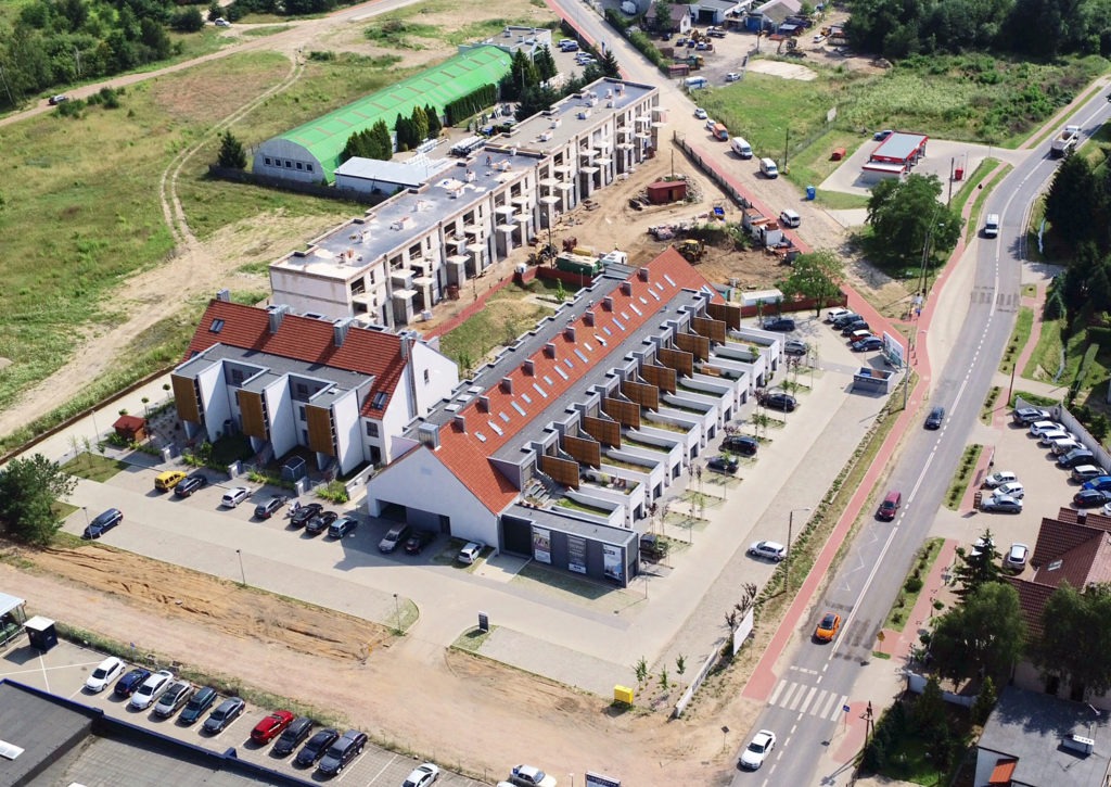 Widok z lotu ptaka na kompleks nowoczesnych domów szeregowych z czerwonymi dachami, częściowo wykończonych, z zaparkowanymi samochodami i otoczeniem w trakcie zagospodarowywania.