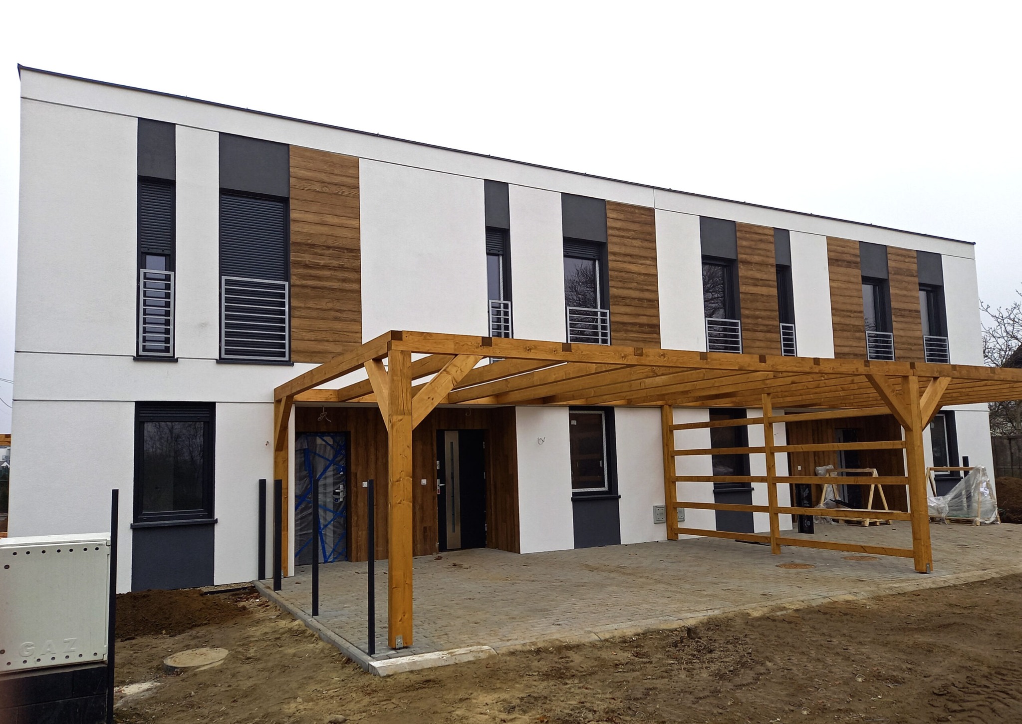 Nowoczesny dom szeregowy z drewnianą pergolą nad miejscem parkingowym, elewacja w kolorze białym i drewnopodobnym, okna z roletami zewnętrznymi.