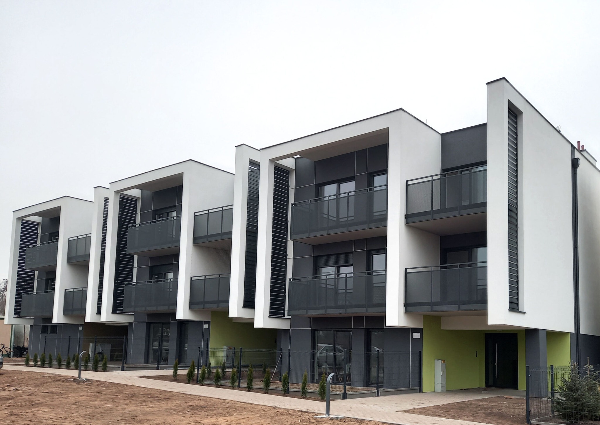 Nowoczesny budynek wielorodzinny z balkonami, wykończony w bieli i szarości, z elementami zieleni w dolnej części, otoczony młodymi drzewkami i chodnikiem z kostki.