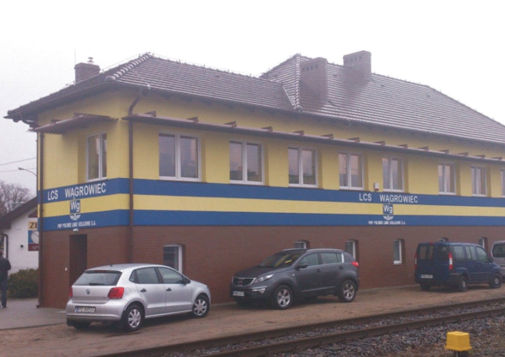 Odnowiony budynek stacji kolejowej z żółtą elewacją, brązowym cokołem i niebieskim pasem, z zaparkowanymi samochodami i torami kolejowymi w kadrze.