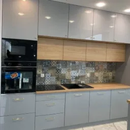 Jasnoszara kuchnia z połyskiem, drewnianymi blatami i szafkami górnymi oraz dekoracyjną mozaiką na ścianie między blatem a szafkami.