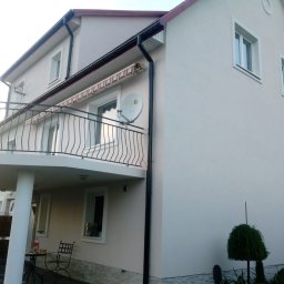 Usługi remontowo budowlane Mateusz Ciacia - Dwupiętrowy dom z balkonem i jasną elewacją, widoczna antena satelitarna, rynny i ozdobne krzewy.
