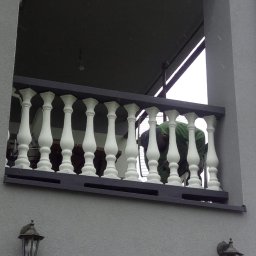 Usługi remontowo budowlane Mateusz Ciacia - Szara elewacja domu z widocznym balkonem z białymi balustradami, osoba w zielonej bluzie pochylona nad balustradą, widoczne lampy elewacyjne.