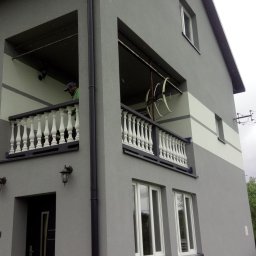 Usługi remontowo budowlane Mateusz Ciacia - Dwukondygnacyjny dom z szarą elewacją, białymi balustradami balkonów i poziomym pasem jaśniejszego koloru, widok z dołu na balkony, gdzie na jednym z nich stoi osoba w zielonej koszuli.