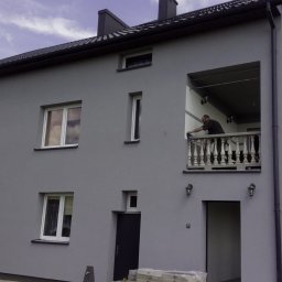 Usługi remontowo budowlane Mateusz Ciacia - Dwupiętrowy dom z szarą elewacją, białe obramowania okien, mężczyzna na balkonie czyści balustradę, oparty o elewację aluminiowy drabina.