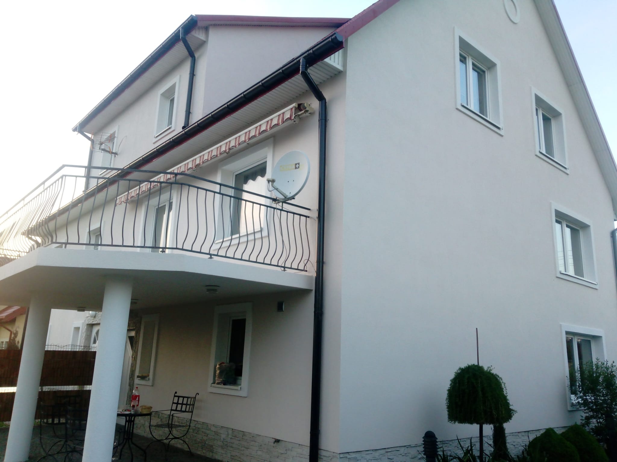 Dwupiętrowy dom z balkonem i jasną elewacją, widoczna antena satelitarna, rynny i ozdobne krzewy.
