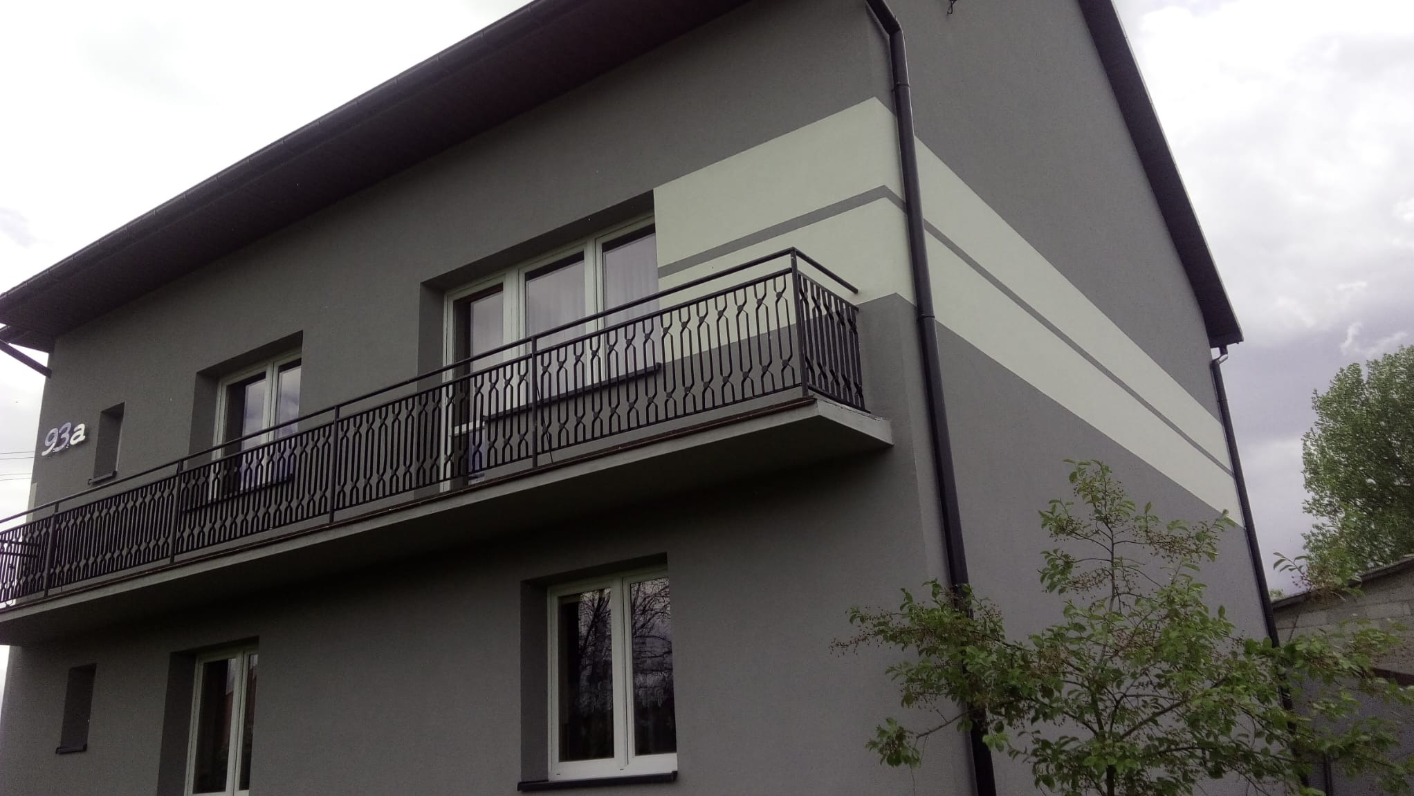 Dwukondygnacyjny dom z szarą elewacją, białymi pasami i balkonem z ozdobną, czarną balustradą, widok z dołu.