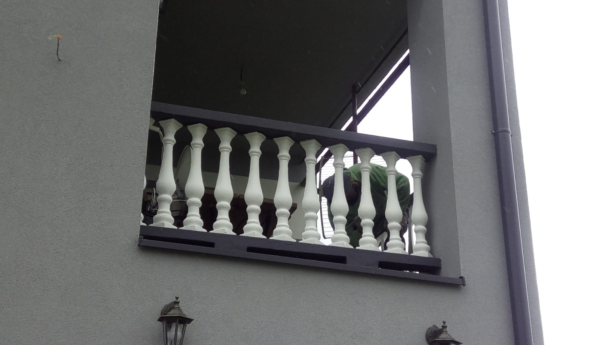 Szara elewacja domu z widocznym balkonem z białymi balustradami, osoba w zielonej bluzie pochylona nad balustradą, widoczne lampy elewacyjne.