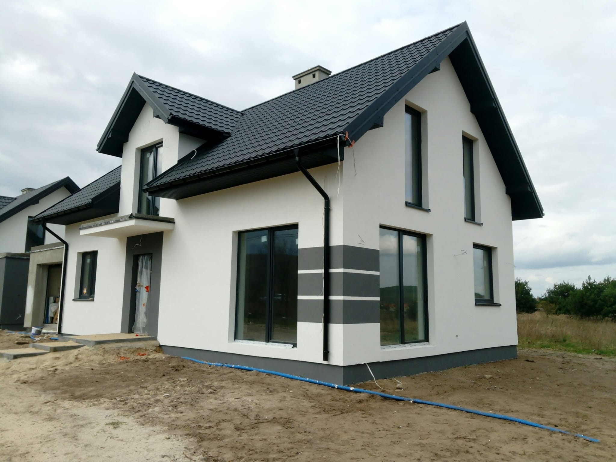 Nowoczesny, dwukondygnacyjny dom z białą elewacją i grafitowym dachem, widoczny w surowym krajobrazie z niezagospodarowanym terenem wokół. Na elewacji widoczne szare pasy dekoracyjne.