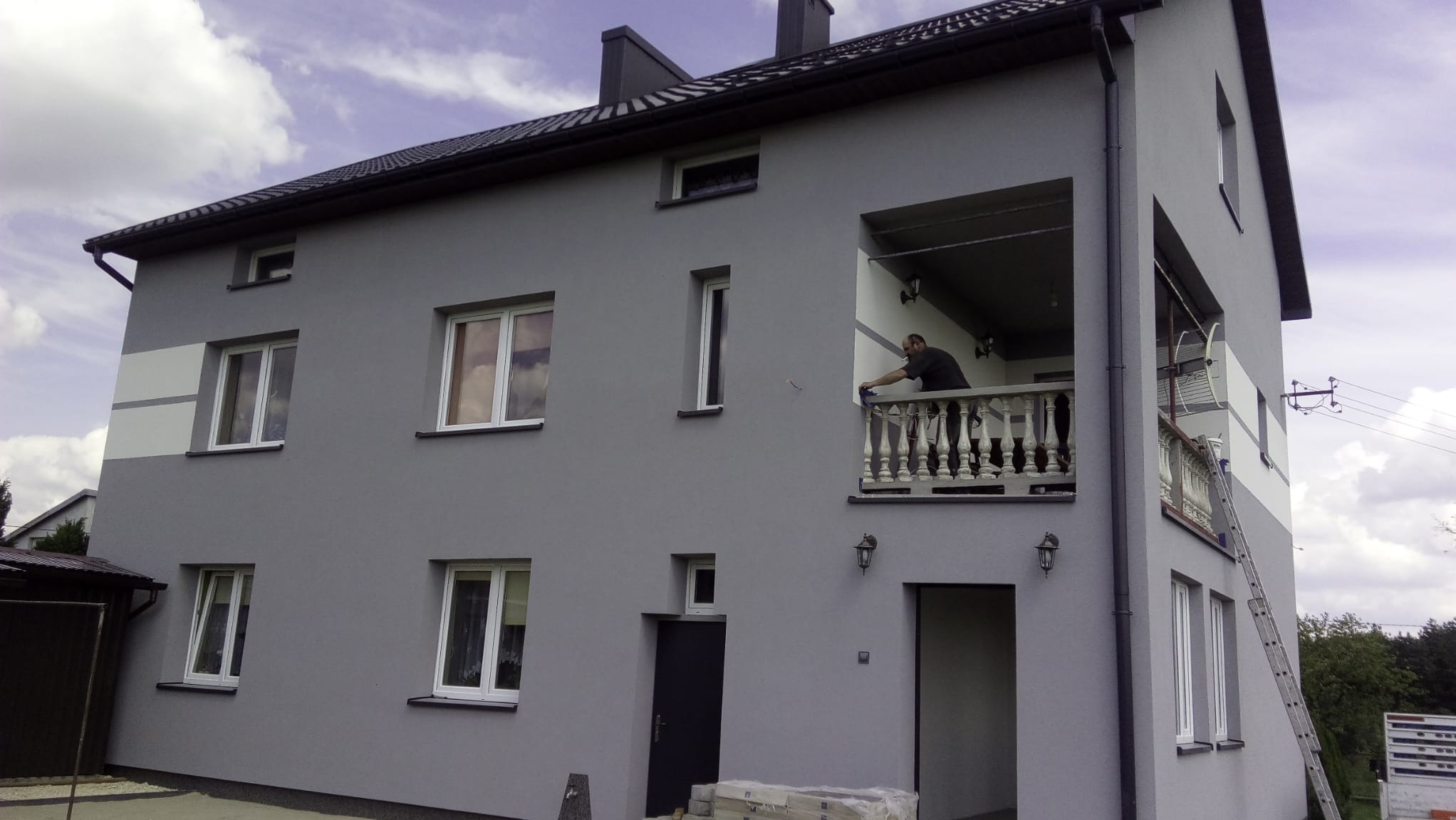 Dwupiętrowy dom z szarą elewacją, białe obramowania okien, mężczyzna na balkonie czyści balustradę, oparty o elewację aluminiowy drabina.