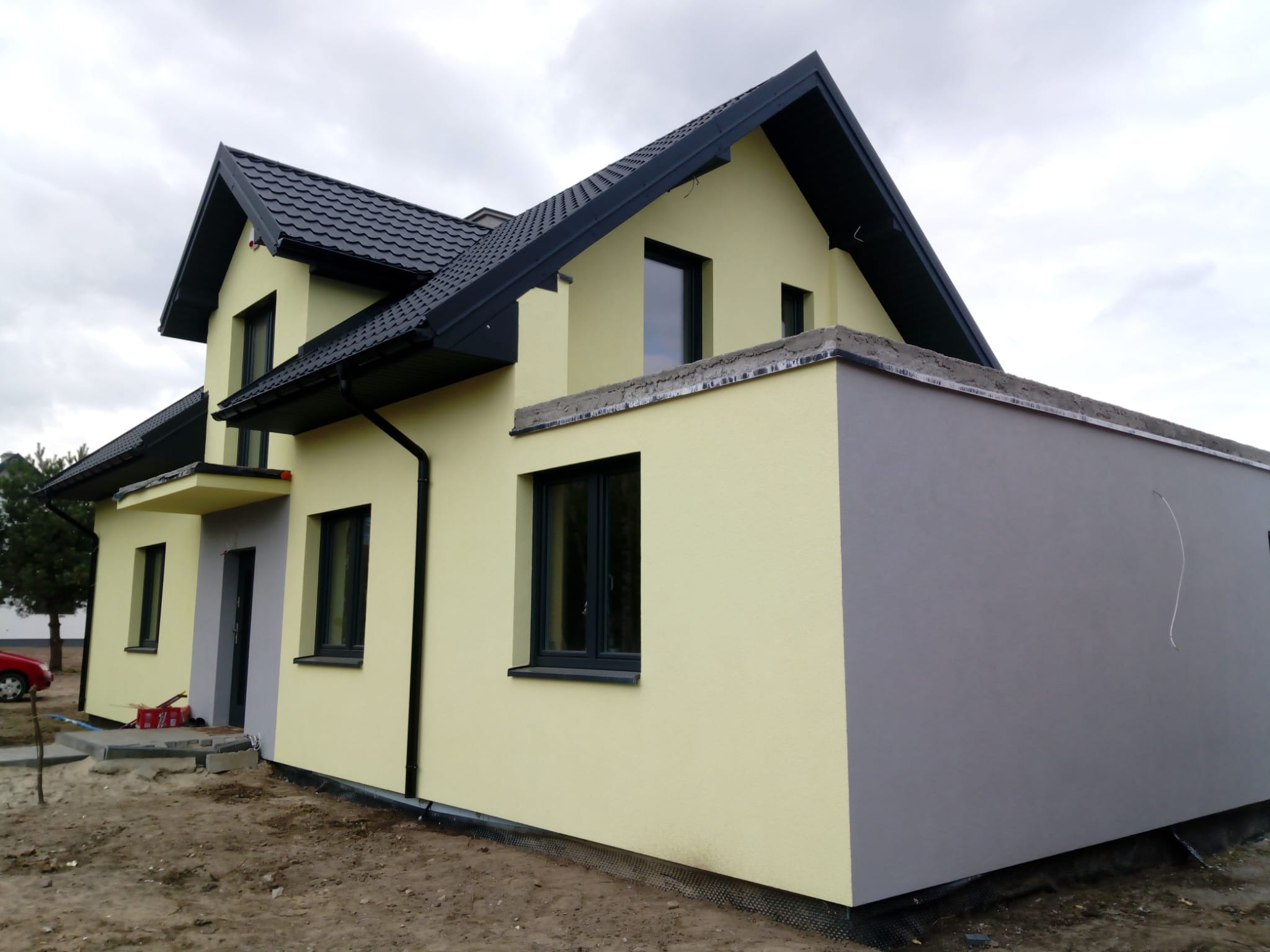 Nowo wybudowany dom jednorodzinny z żółtą elewacją i grafitowym dachem, widoczne fragmenty niezabudowanej działki i podłoże pod kostkę brukową.