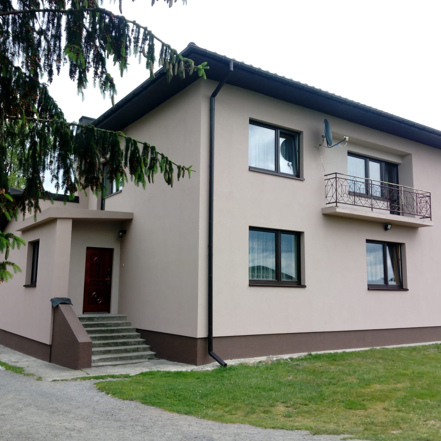 Dwukondygnacyjny dom z beżową elewacją, brązowymi detalami, balkonem z ozdobną balustradą i anteną satelitarną na ścianie, widziany z zewnątrz w pochmurny dzień.