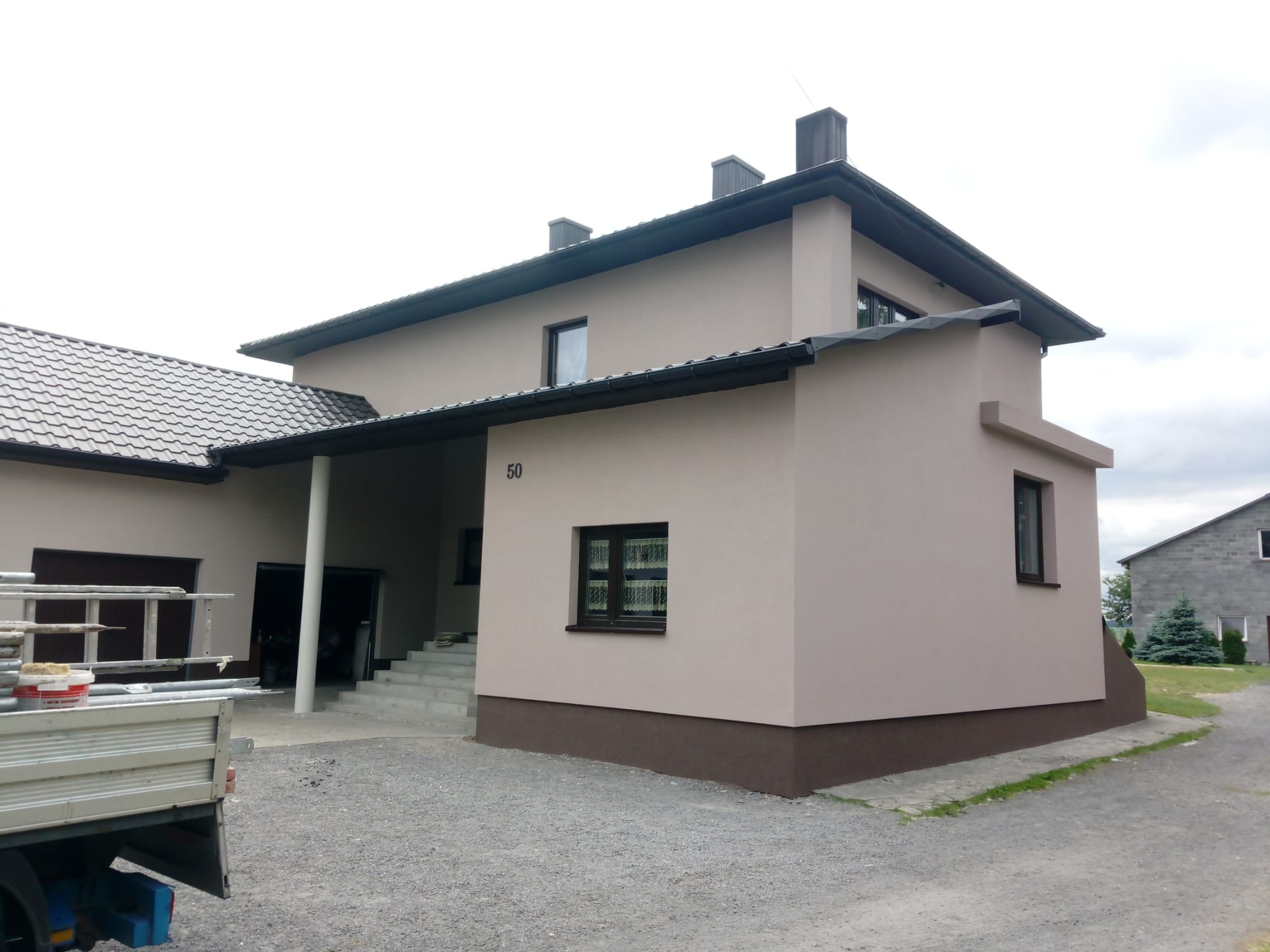 Wykończony dom z beżową elewacją, ciemnobrązowym cokołem i grafitowym dachem, z widoczną ciężarówką z akcesoriami malarskimi i drabiną, sugerującą prace wykończeniowe.