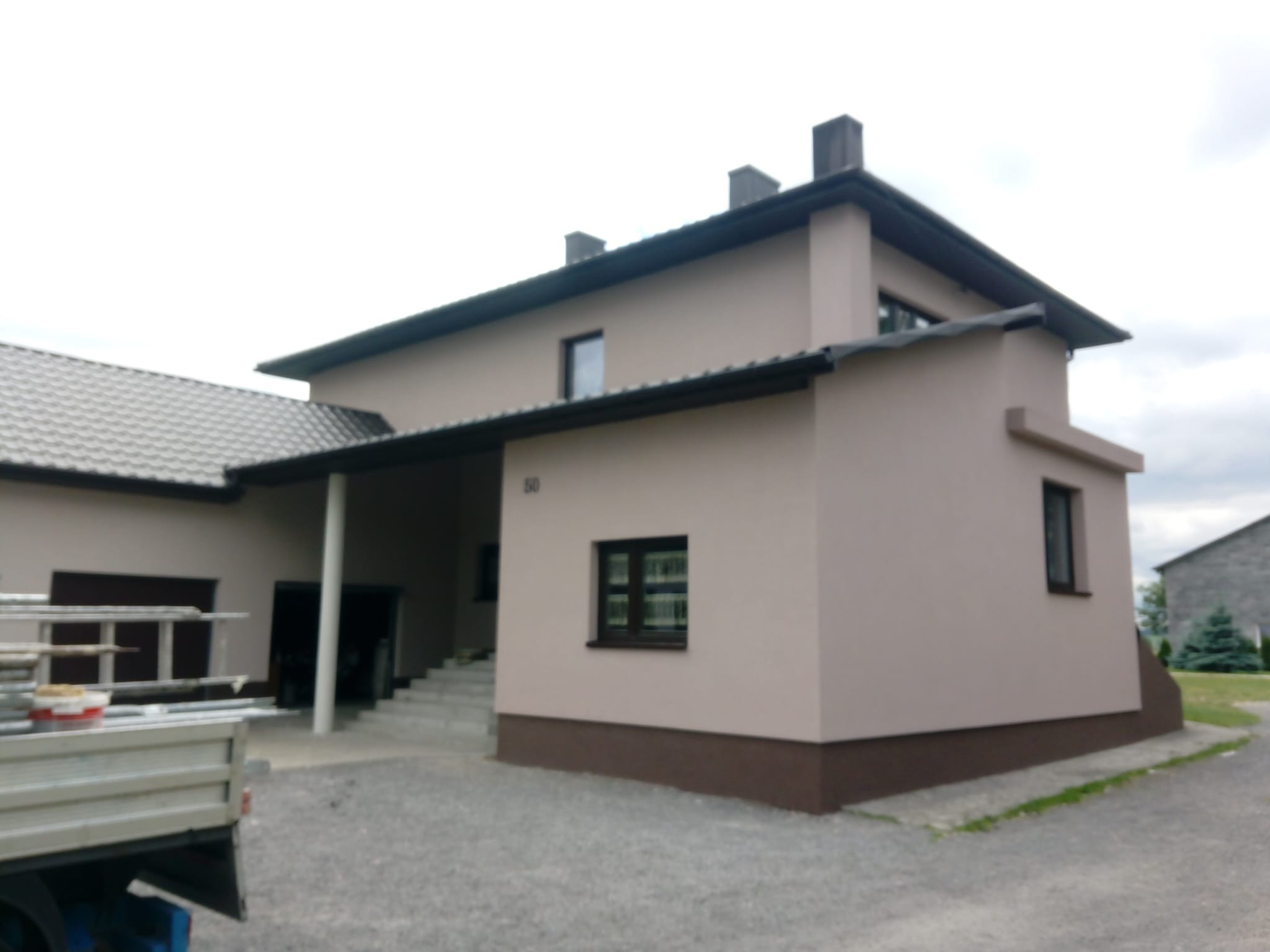 Dwukondygnacyjny dom z jasną elewacją i ciemnobrązowym cokołem, widok z boku na podjazd z zaparkowanym samochodem dostawczym z drabiną i pojemnikami z farbą.