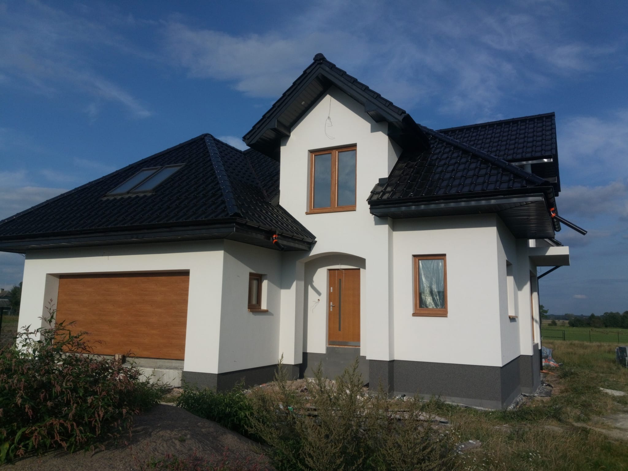 Nowoczesny dom jednorodzinny z ciemną dachówką, jasną elewacją i drewnianą bramą garażową w słoneczny dzień.