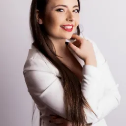 Portret uśmiechniętej kobiety w białym żakiecie, z ciemnymi włosami opadającymi na ramię i czerwonymi ustami, pozującej na jasnym tle z ręką pod brodą.