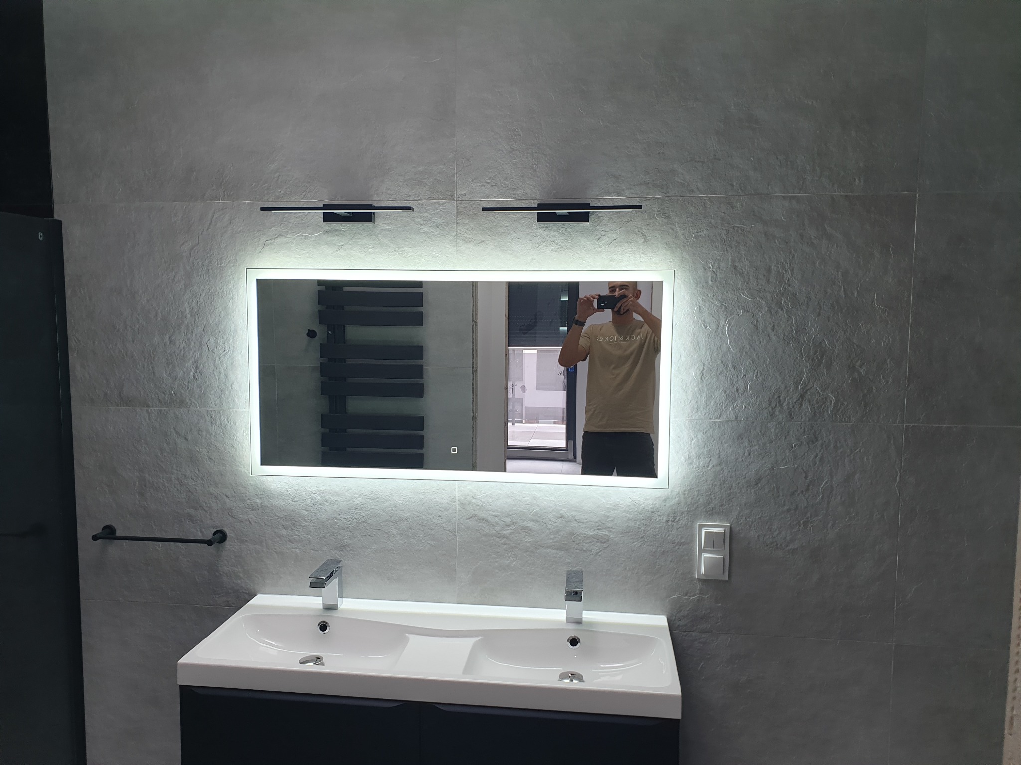 Nowoczesna łazienka z szarymi płytkami imitującymi kamień, podświetlanym lustrem i podwójną umywalką, widoczne odbicie fotografa w lustrze.