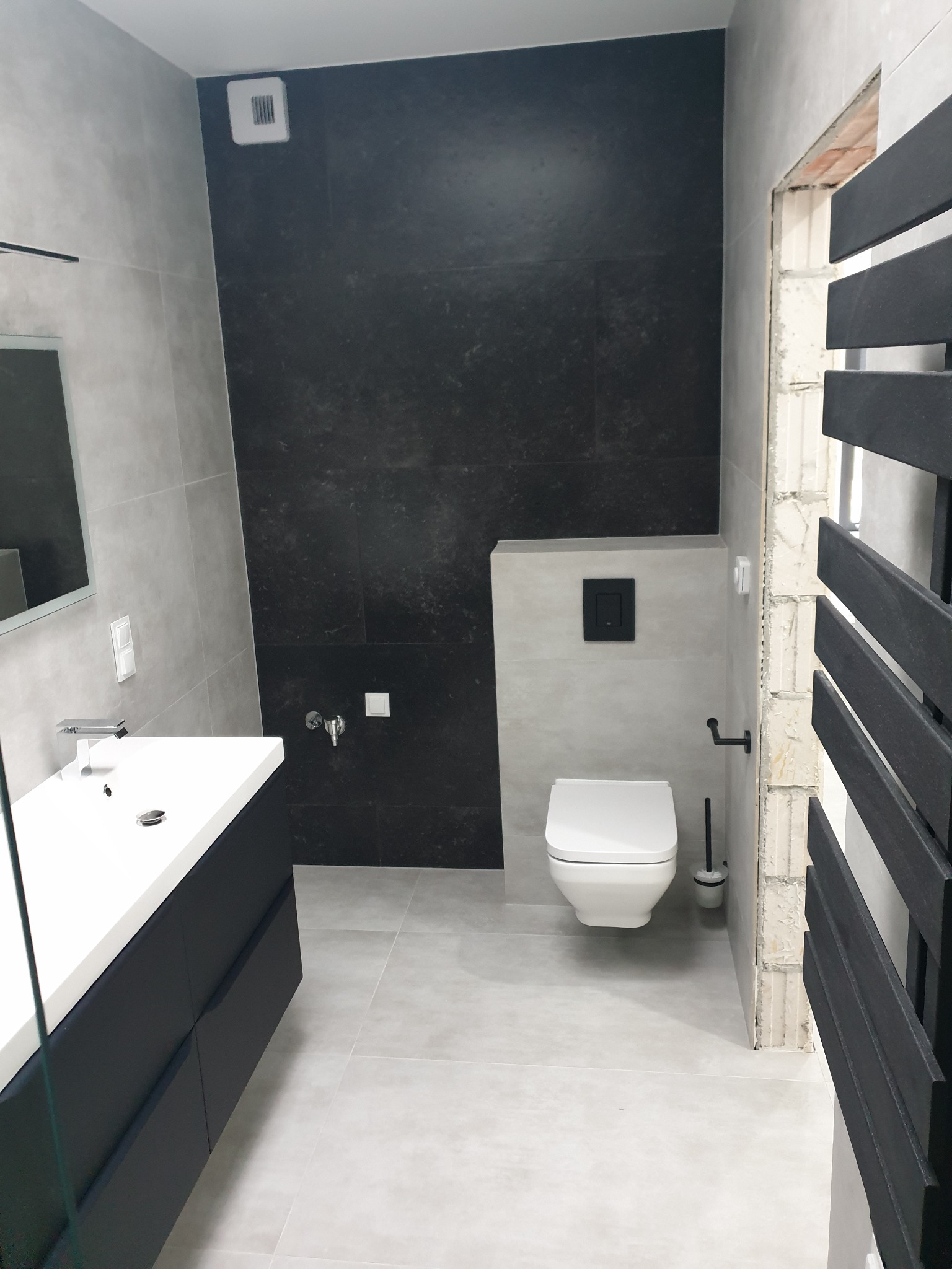 Nowoczesna łazienka z szarymi i czarnymi płytkami, wisząca toaleta z czarnym przyciskiem, umywalka z szafką w kolorze antracytowym, grzejnik drabinkowy w kolorze czarnym i fragment ściany w trakcie...