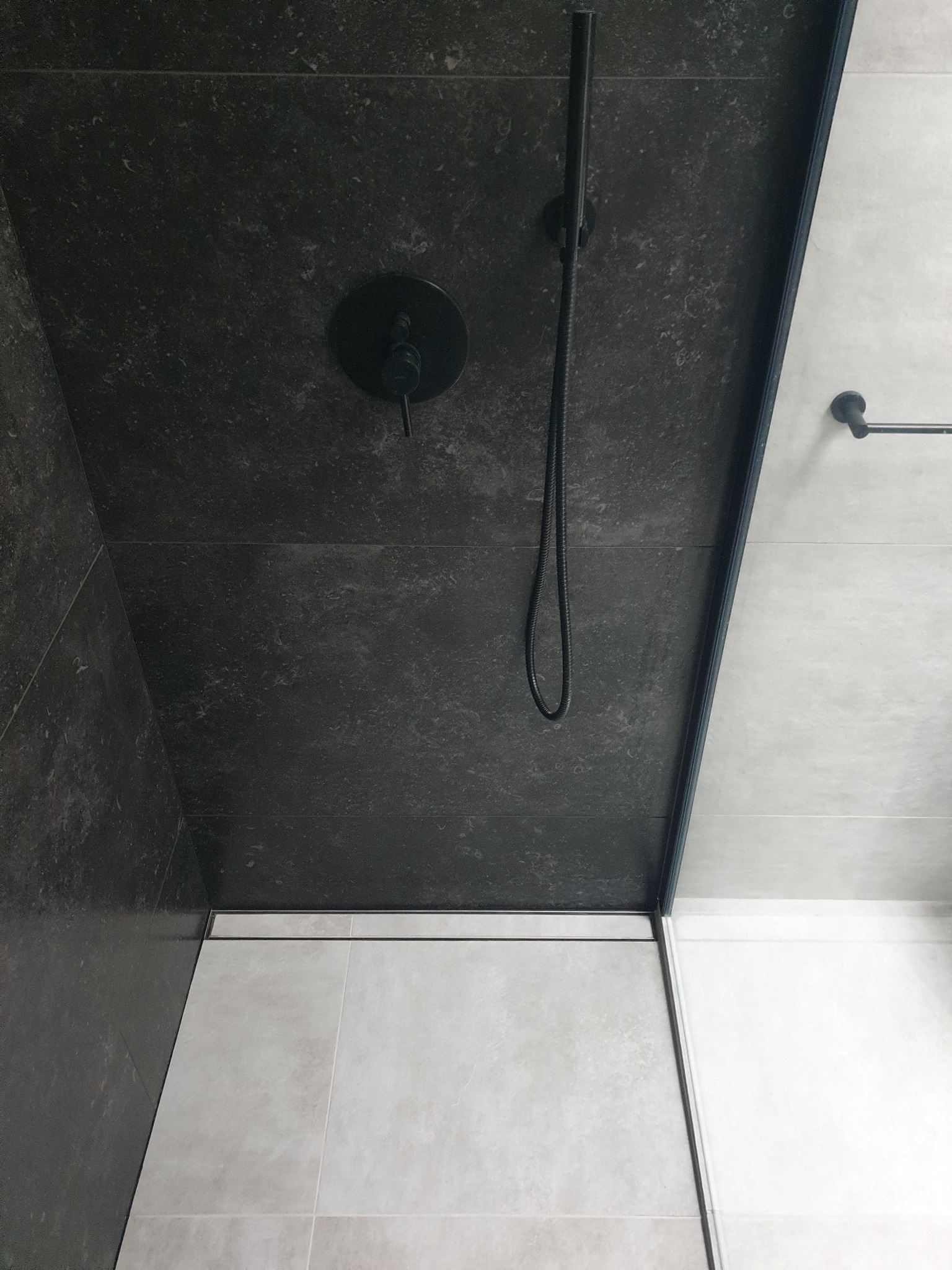 Nowoczesna kabina prysznicowa z ciemnoszarą płytką na ścianach i suficie, czarna armatura, odpływ liniowy i jasne płytki podłogowe.