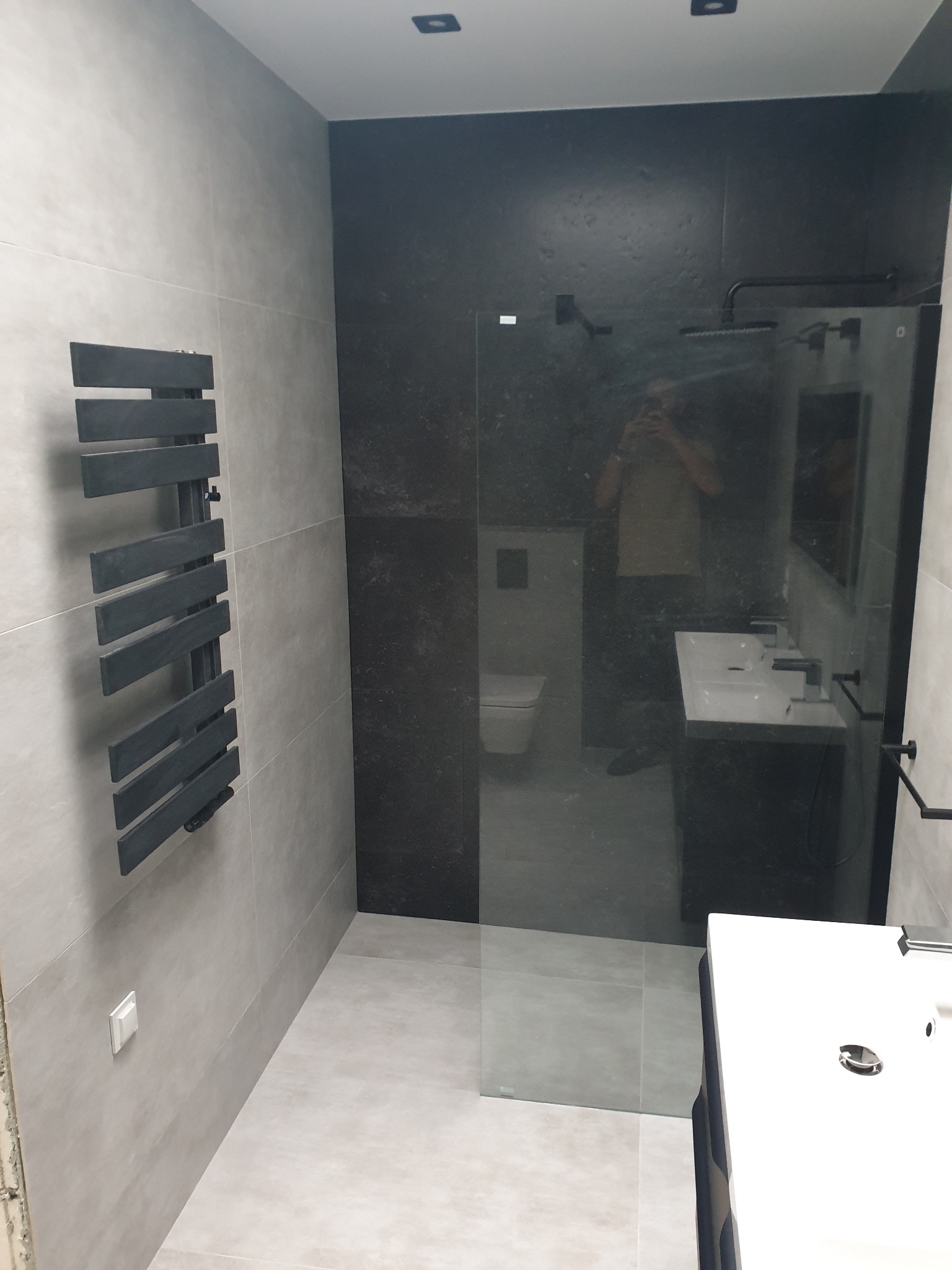 Nowoczesna łazienka z kabiną prysznicową typu walk-in, grzejnikiem drabinkowym w kolorze antracytowym, umywalką nablatową i czarną ścianą akcentującą.