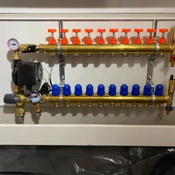 Rozdzielacz do instalacji grzewczej z dziesięcioma obwodami, z czerwonymi i niebieskimi zaworami, zamontowany w białej szafce, widoczne manometry i pompa obiegowa.