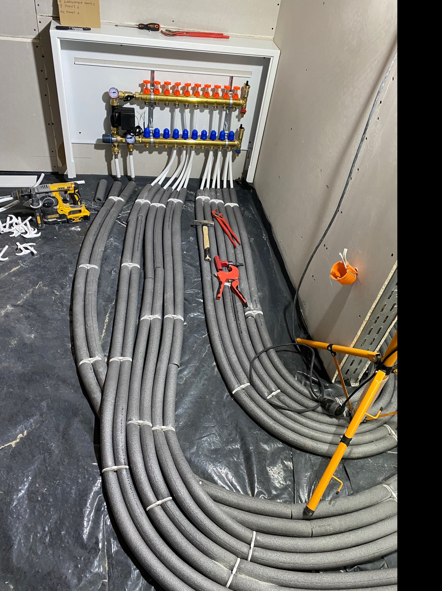 Rozdzielacz ogrzewania podłogowego z podłączonymi, zaizolowanymi rurami, narzędzia pracy, skrzynka z notatkami na ścianie w surowym wnętrzu.