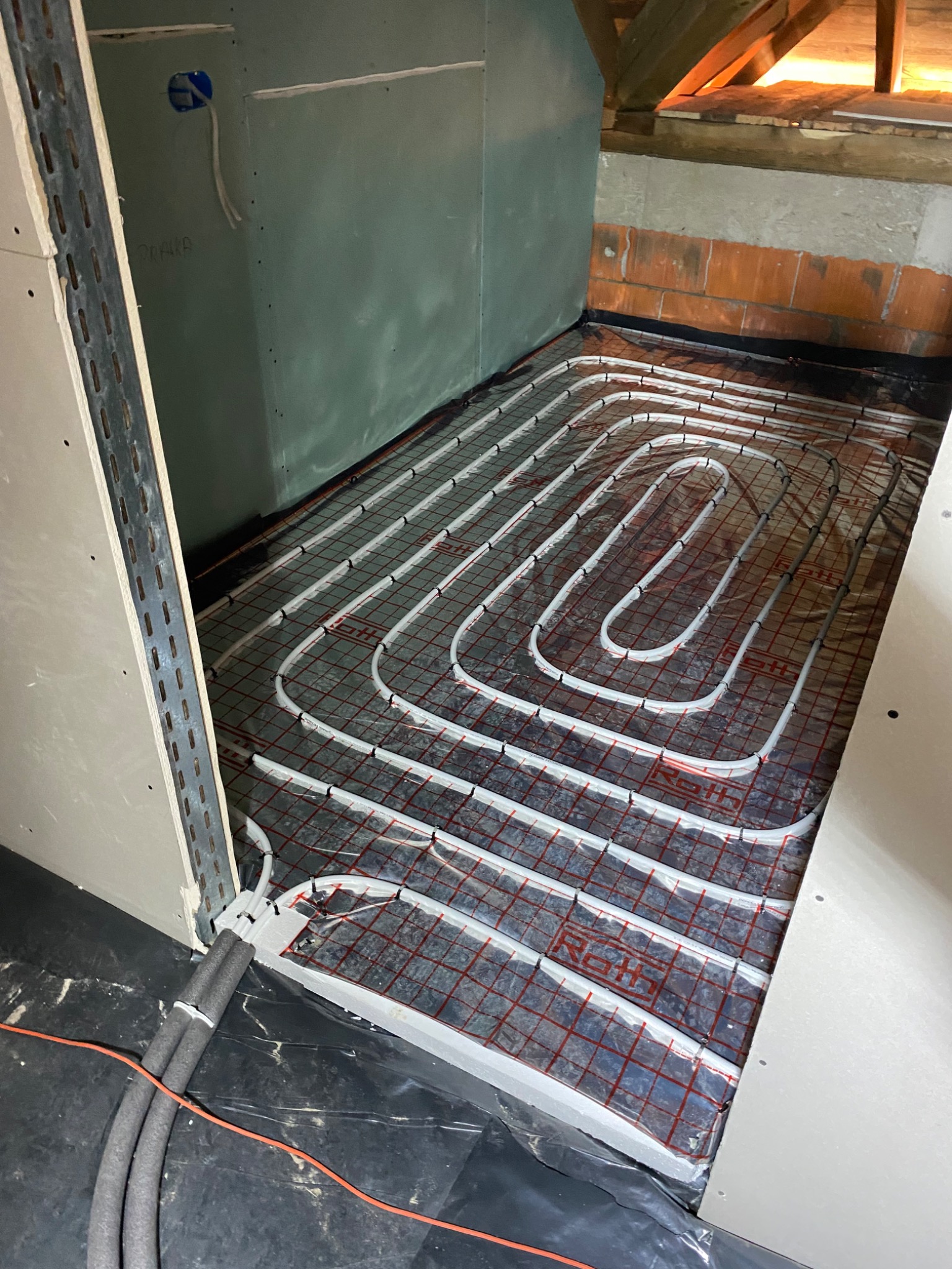 Układanie białych rurek systemu grzewczego na czerwonej siatce w pomieszczeniu na poddaszu, widoczne izolowane rury doprowadzające i fragment ściany z płyt gipsowych.