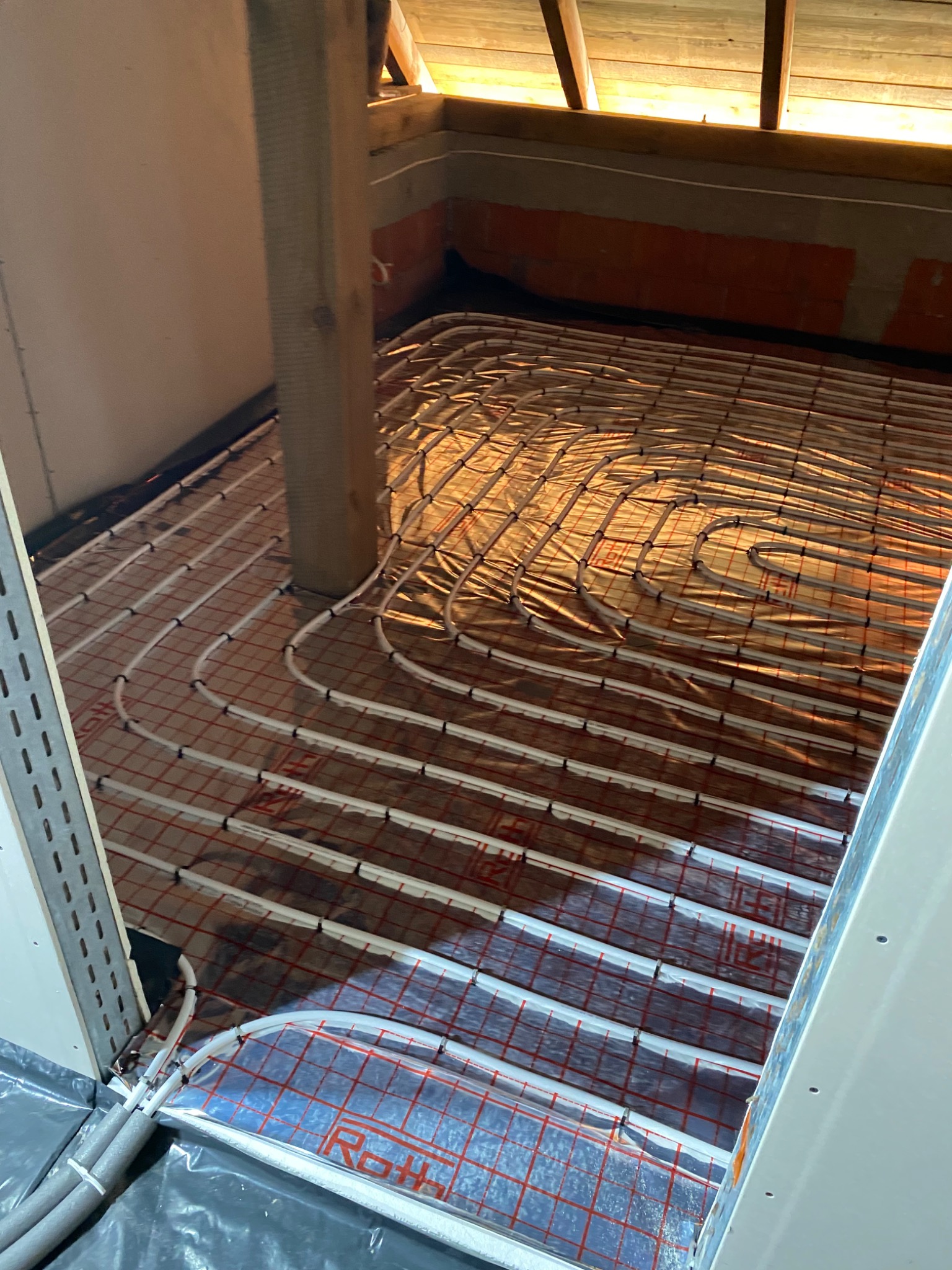 Białe rurki systemu rozprowadzenia ciepła na folii z czerwoną siatką, zamontowane w poddaszu, widok z góry pod kątem.