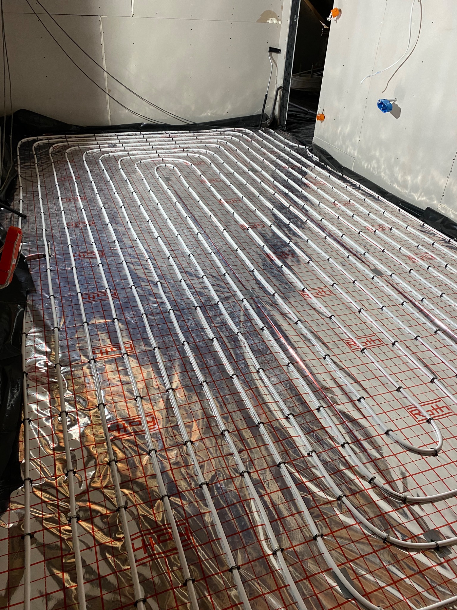 Rozłożone rury ogrzewania podłogowego na srebrnej folii z czerwoną siatką, widoczne czarne mocowania rur, ściany w surowym stanie.