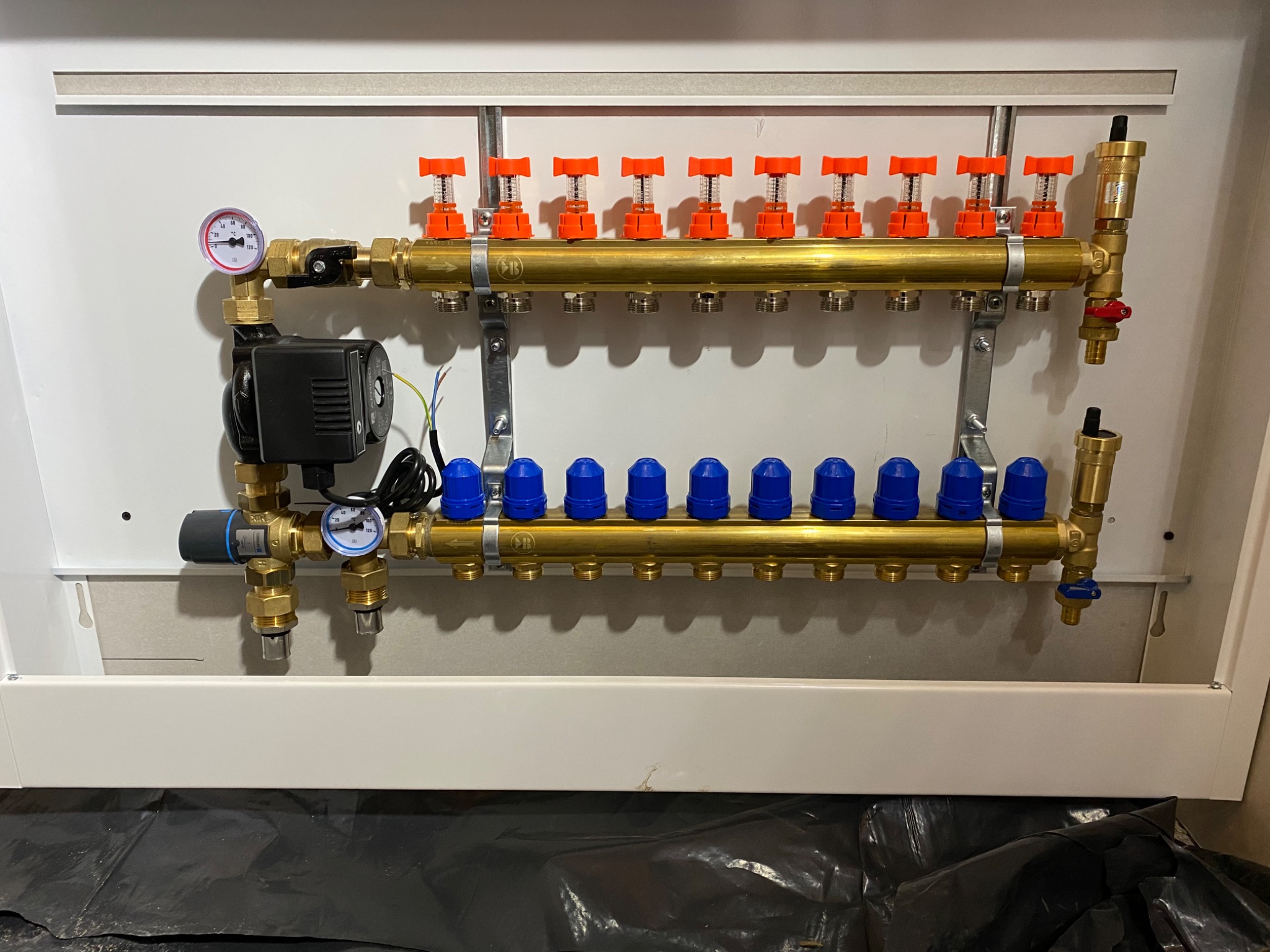 Rozdzielacz do instalacji grzewczej z dziesięcioma obwodami, z czerwonymi i niebieskimi zaworami, zamontowany w białej szafce, widoczne manometry i pompa obiegowa.