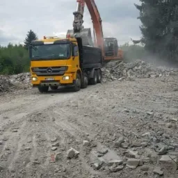 Żółta ciężarówka Mercedes Benz Actros z czarną naczepą, ładowana gruzem przez pomarańczową koparkę na terenie po rozbiórce budynku, w tle drzewa i pochmurne niebo.