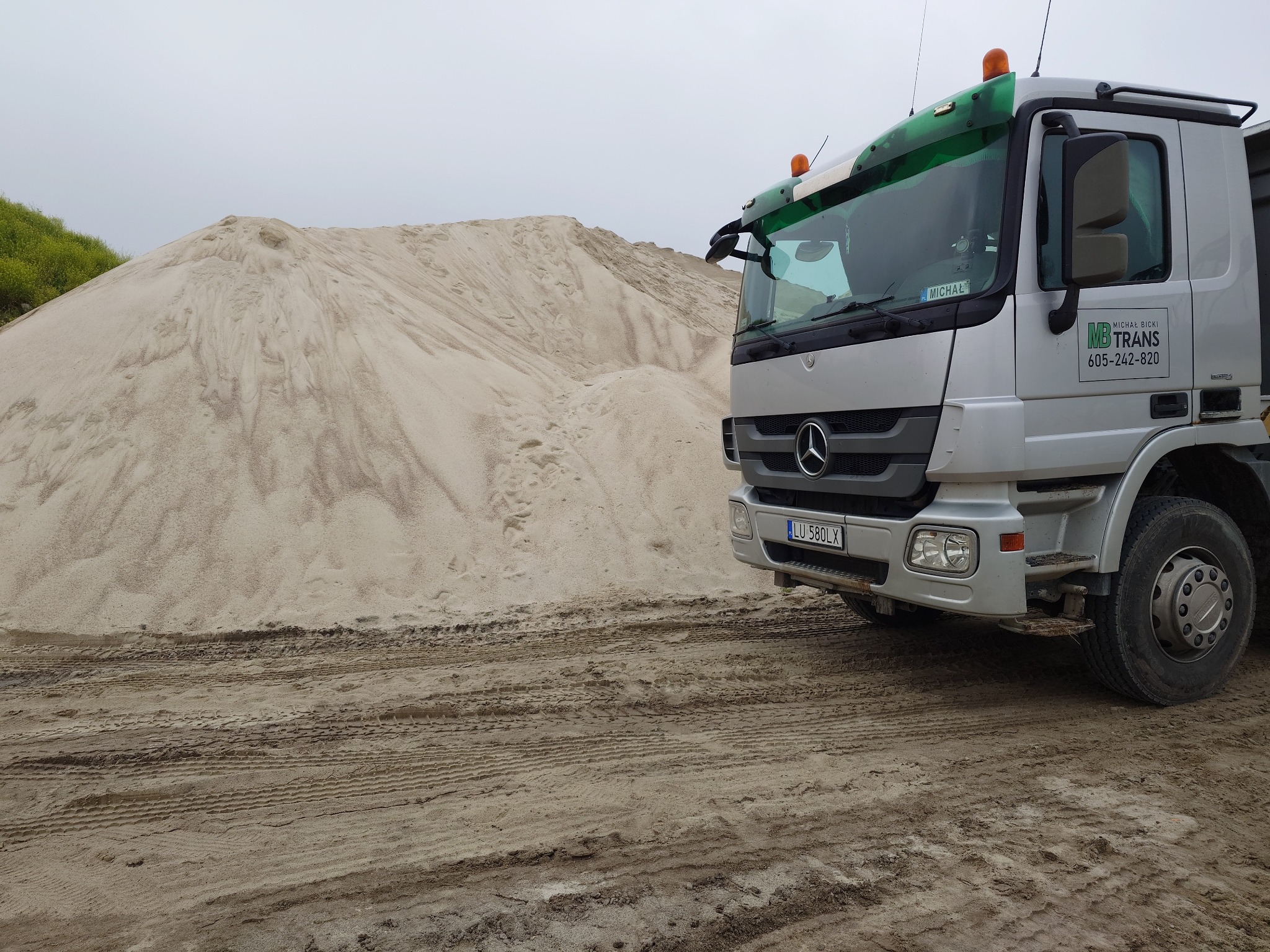 Srebrna ciężarówka z logo MB Trans zaparkowana obok sporej pryzmy piasku na placu budowy, widoczne ślady opon na ziemi.