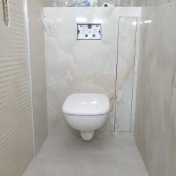 Dom-Jura - Wykończona toaleta podwieszana z beżowymi płytkami imitującymi marmur, widoczny stelaż podtynkowy i rewizja. Minimalistyczny design, ciepłe oświetlenie.