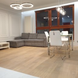 Dom-Jura - Jasne panele podłogowe w salonie z szarą sofą narożną, białymi krzesłami i stołem ze szklanym blatem. Okno z drewnianą ramą i nowoczesne lampy.