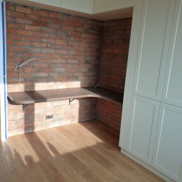 Dom-Jura - Kącik z biurkiem z blatem w kolorze drewna, wspartym na metalowych wspornikach, na tle ściany z czerwonej cegły i białej zabudowy meblowej. Widoczne przewody elektryczne.