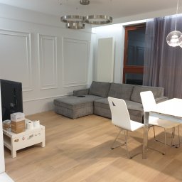 Dom-Jura - Jasne panele podłogowe w salonie z szarą sofą i białymi krzesłami. Ściany zdobią dekoracyjne listwy. Nowoczesne oświetlenie sufitowe.