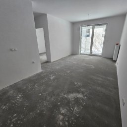Dom-Jura - Jasne pomieszczenie z białymi, gładkimi ścianami i surową, betonową podłogą z pyłem, przygotowane do dalszych prac. Widoczne podwójne włączniki światła, nowoczesne drzwi balkonowe i grzejnik.