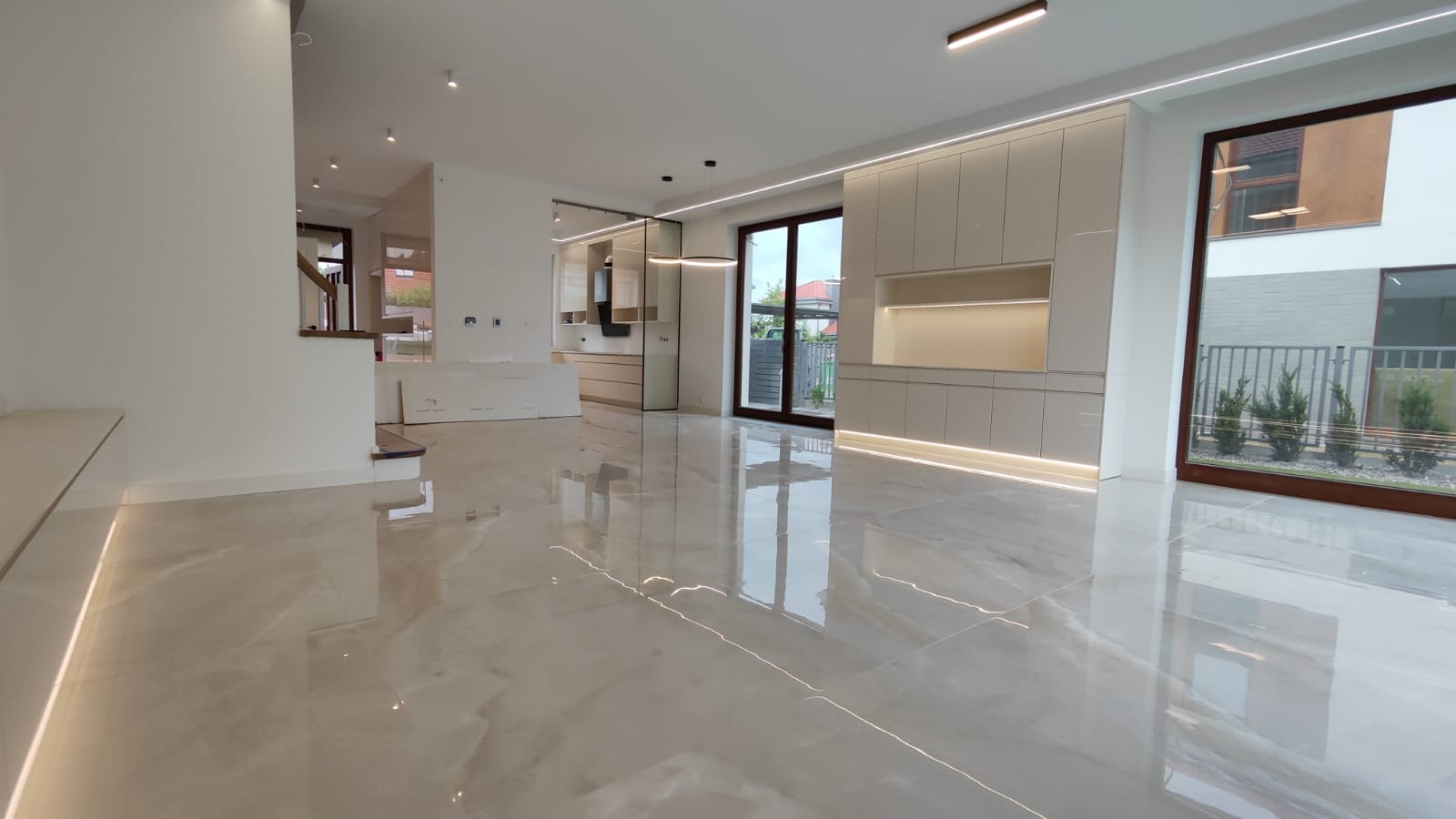 Przestronne, jasne wnętrze po remoncie z błyszczącą, marmurkową podłogą, nowoczesną zabudową kuchenną z podświetleniem LED i widokiem na ogród przez duże okno.